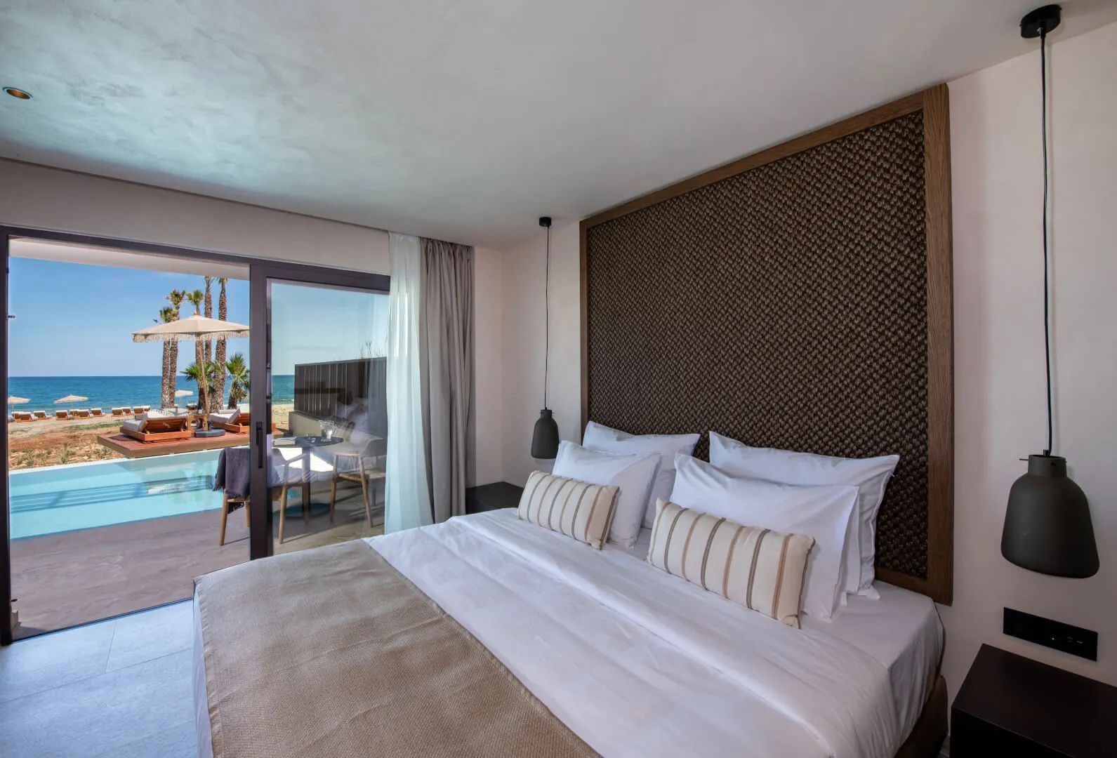 Doppelbett im Schlafbereich mit Blick durch die Glasflügeltür auf die Terrasse, den Pool und das Meer.