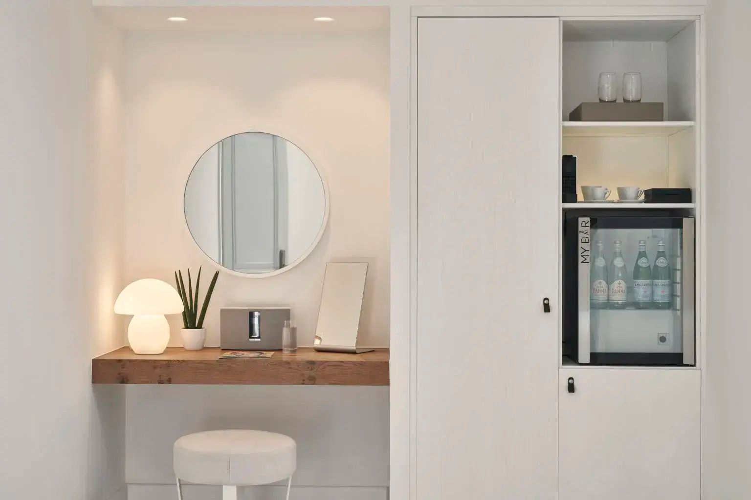 Schminktisch mit rundem Wandspiegel, Tischleuchte, Hocker und angrenzendem weißem Einbauschrank.