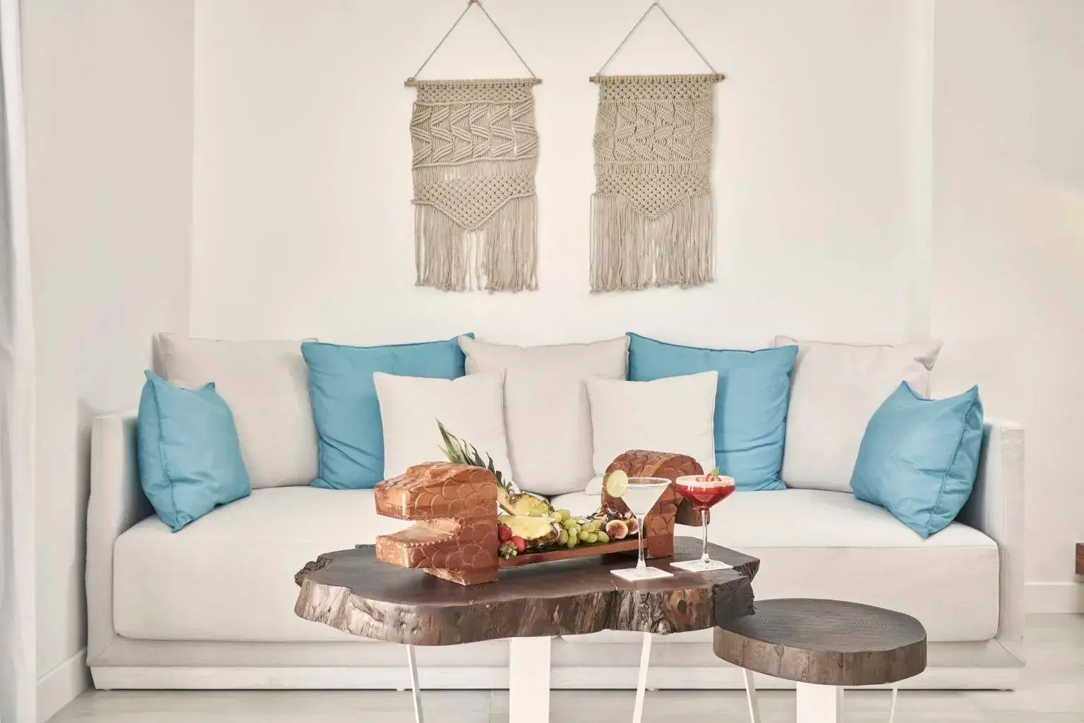 Wohnbereich mit weißem Sofa, hellblauen Kissen, Holztisch und Wanddekoration aus Makramee.
