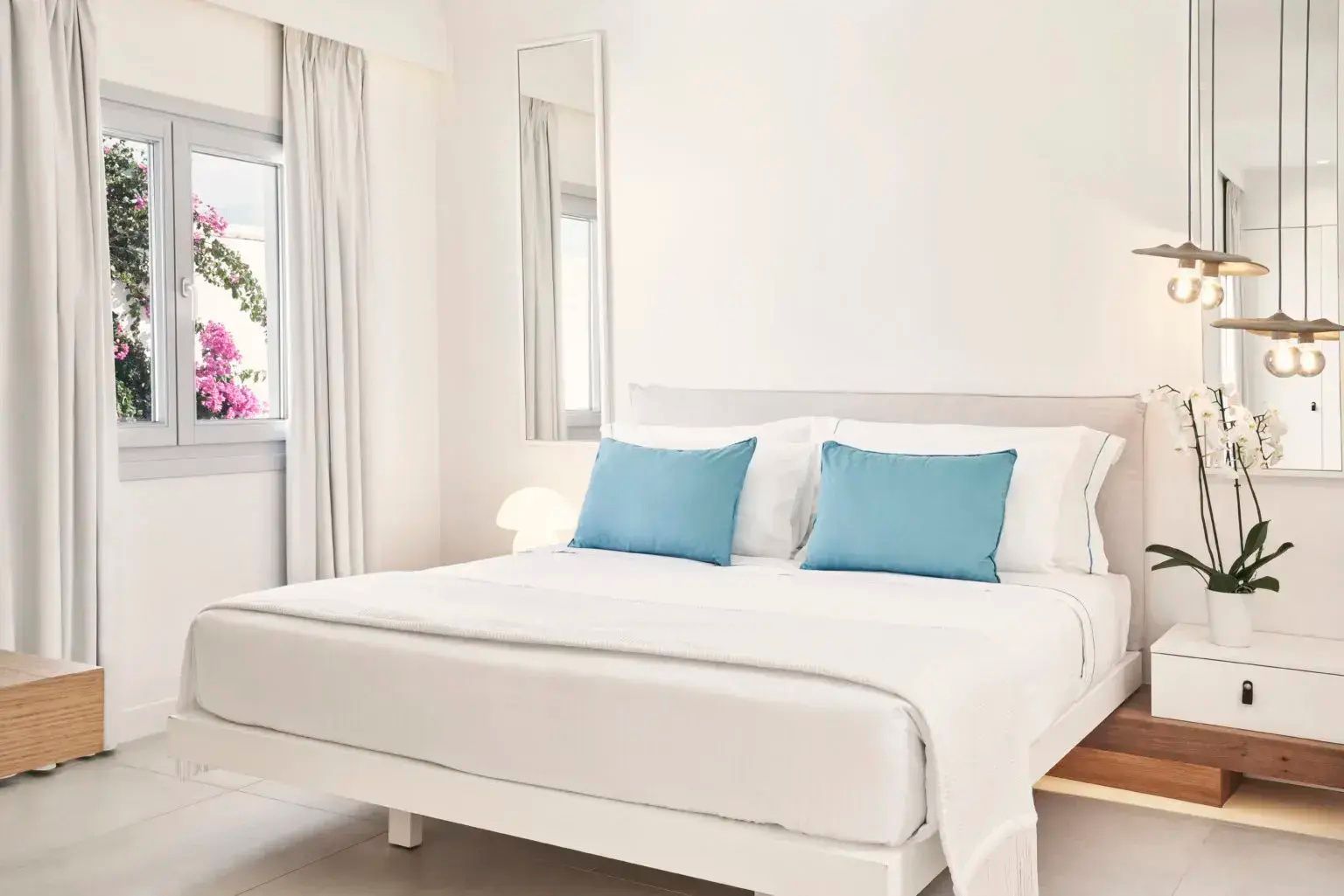 Weißes Doppelbett mit blauen Kissen in einem hell gestalteten Hotelzimmer.
