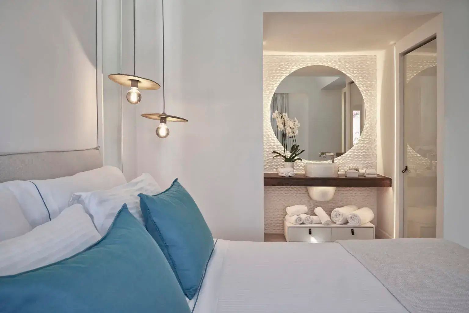 Blick vom Bett mit blauen Kissen auf den angrenzenden Waschtisch mit rundem Spiegel.