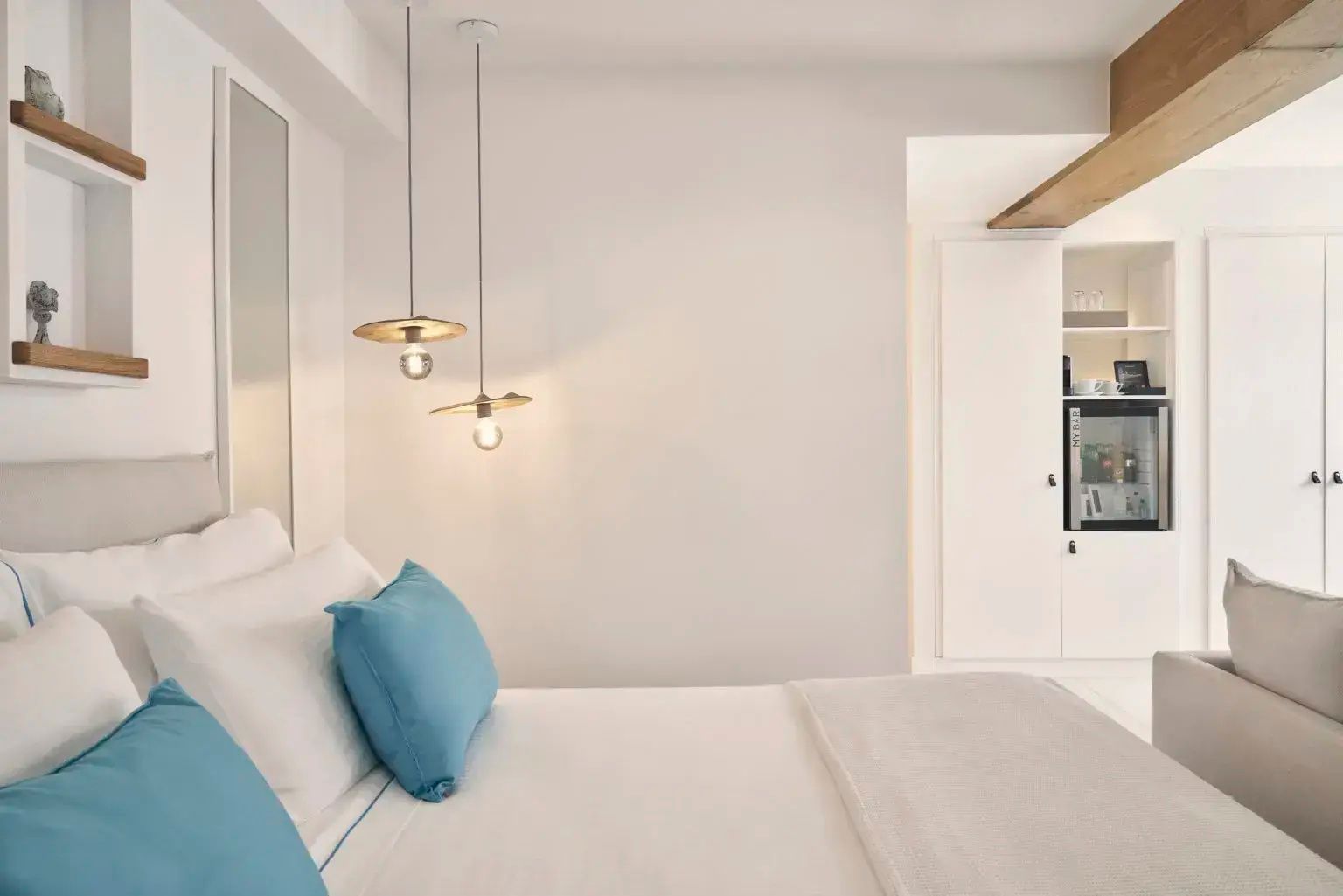 Doppelbett mit weißen Decken, blauen Kissen und hängenden Leuchten in einem hellen Hotelzimmer.