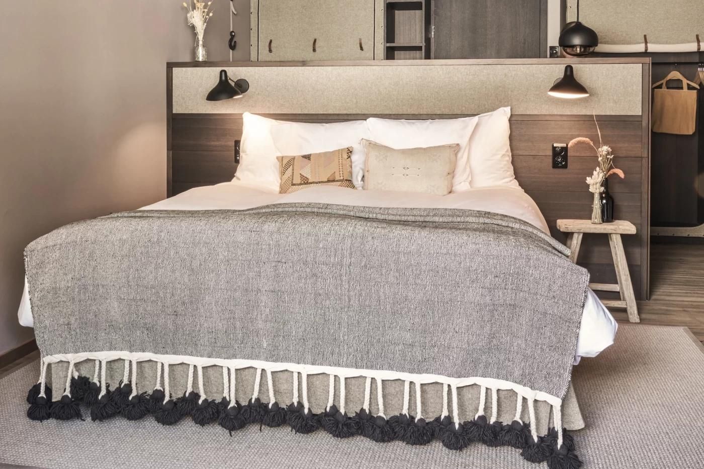 Doppelbett im Hotelzimmer mit grauer Fransen-Tagesdecke, weichen Kissen und Holzkopfteil.