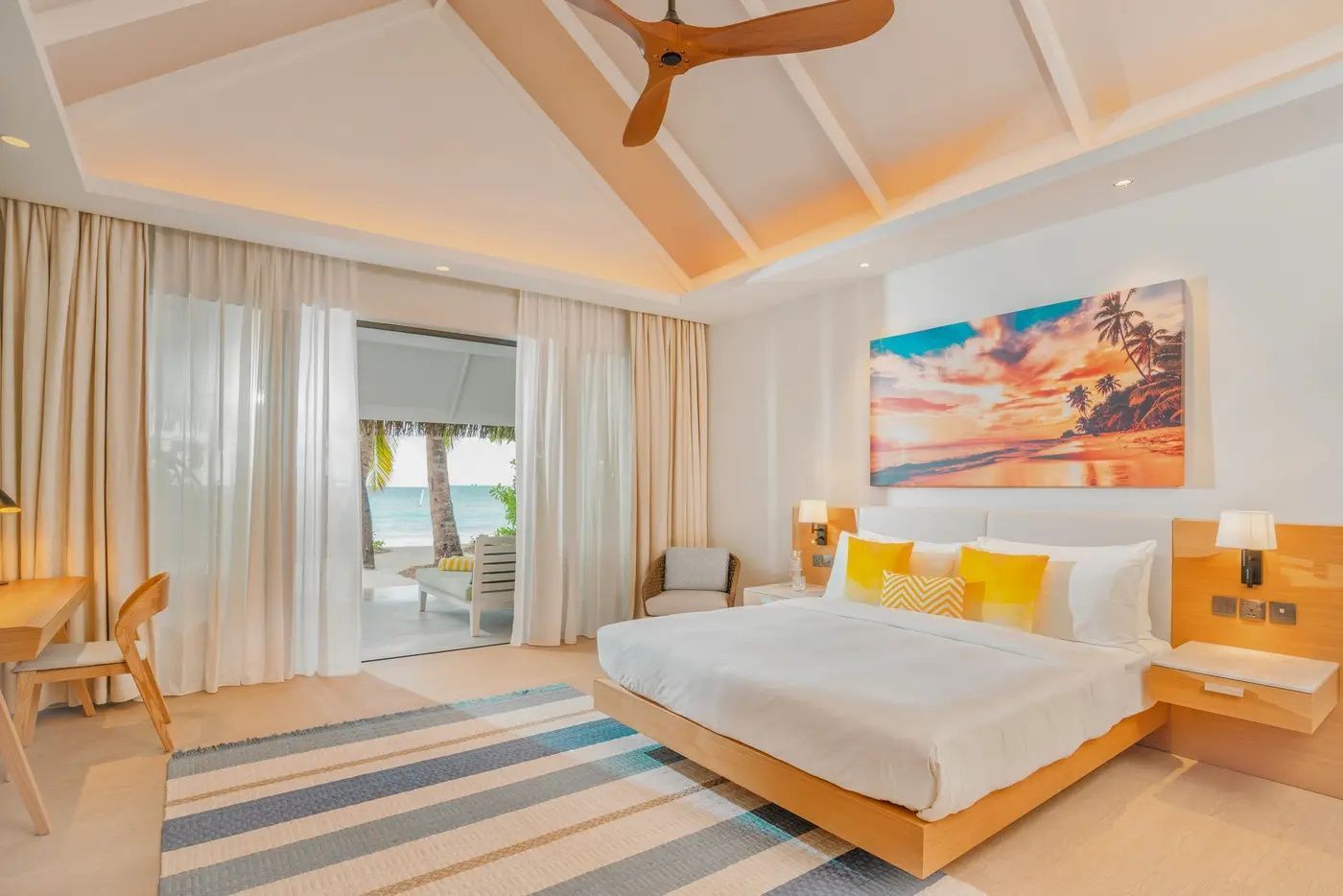 Helles Schlafzimmer mit Doppelbett, gelben Kissen, Deckenventilator und Blick auf den Strand.