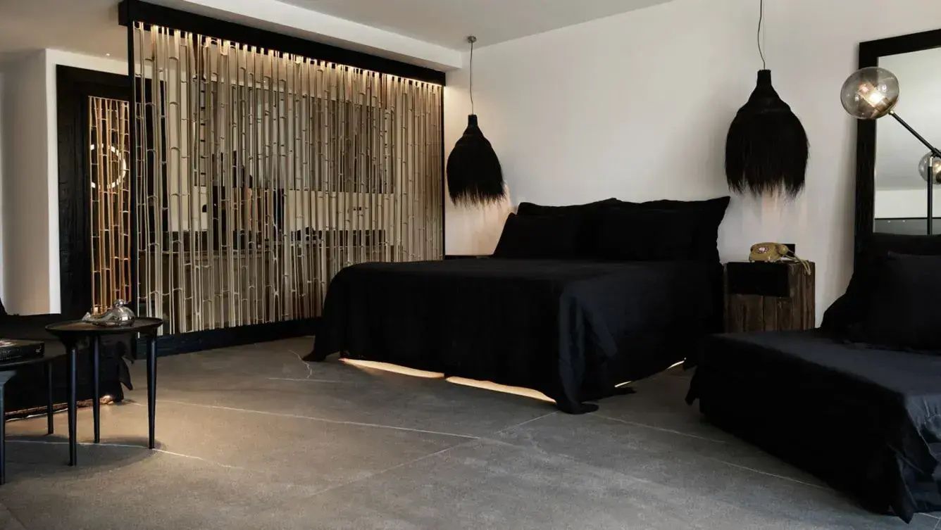 Hotelzimmer mit schwarzem Doppelbett, Holztrennwand und dunklem Steinboden.