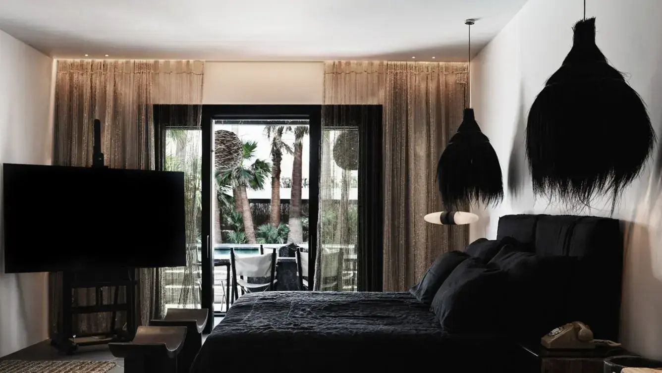 Dunkles Doppelbett im Hotelzimmer mit Blick durch die Glasfront auf Palmen im Außenbereich.