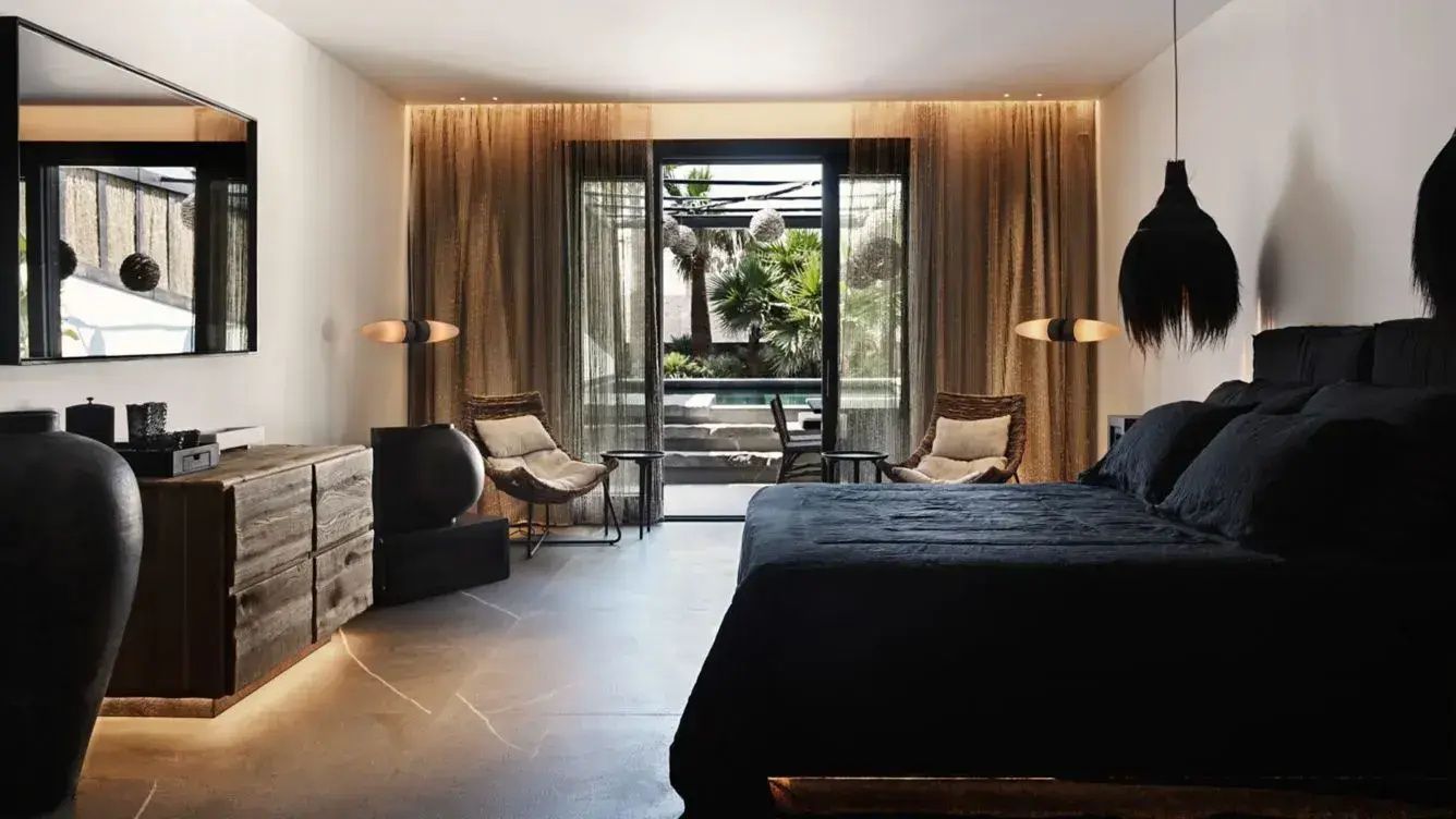 Schlafzimmer mit dunkler Bettwäsche und Vorhängen vor einer Terrasse mit Palmen.