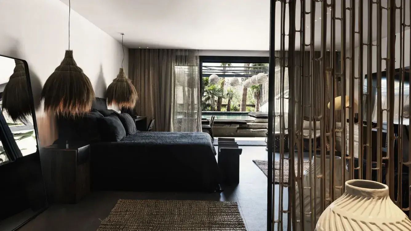Modernes Hotelzimmer mit dunkel bezogenem Doppelbett, Flechtlampe und Blick auf die Terrasse.