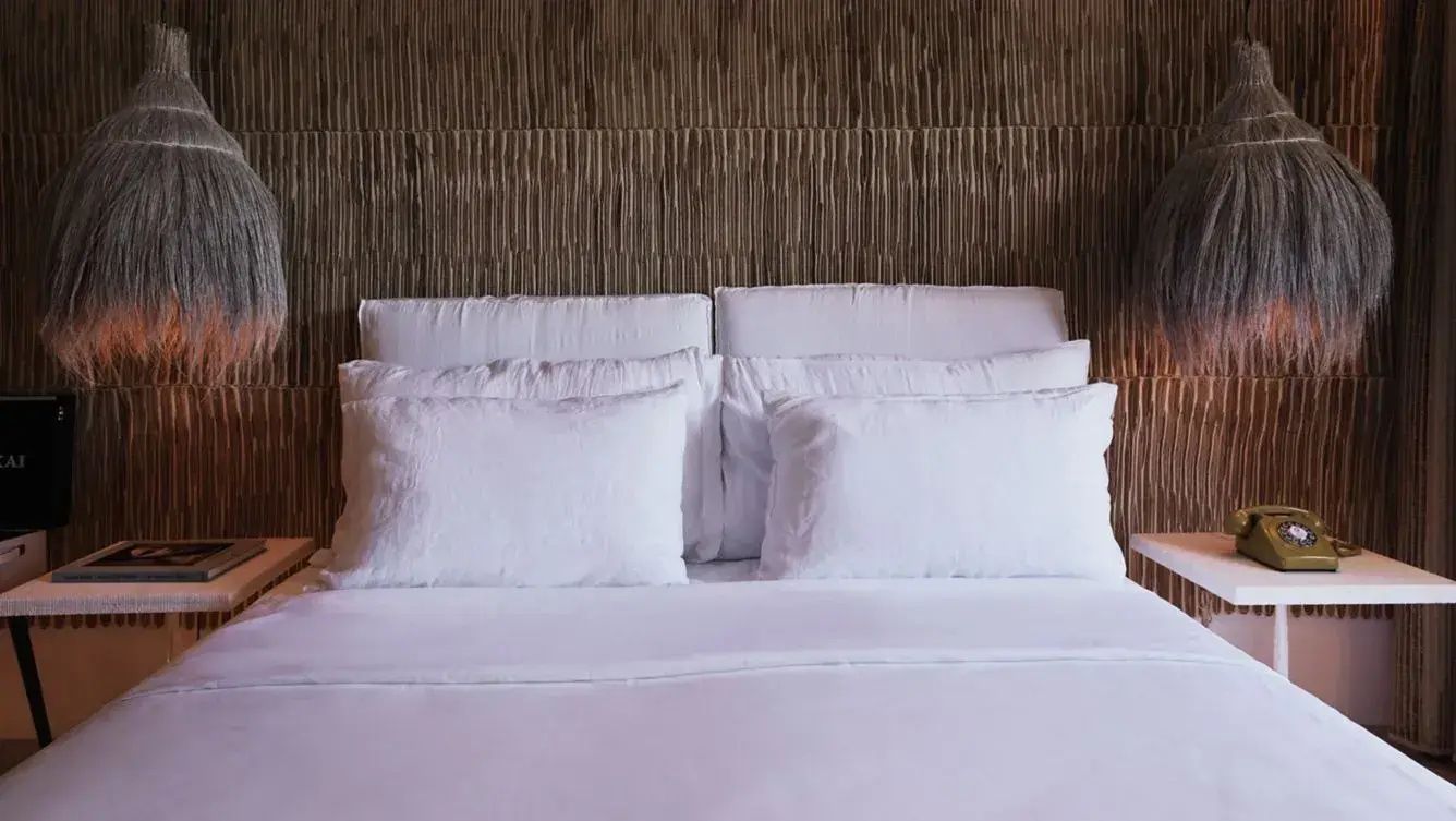 Weiß bezogenes Doppelbett mit mehreren Kissen vor einer strukturierten Holzrückwand.