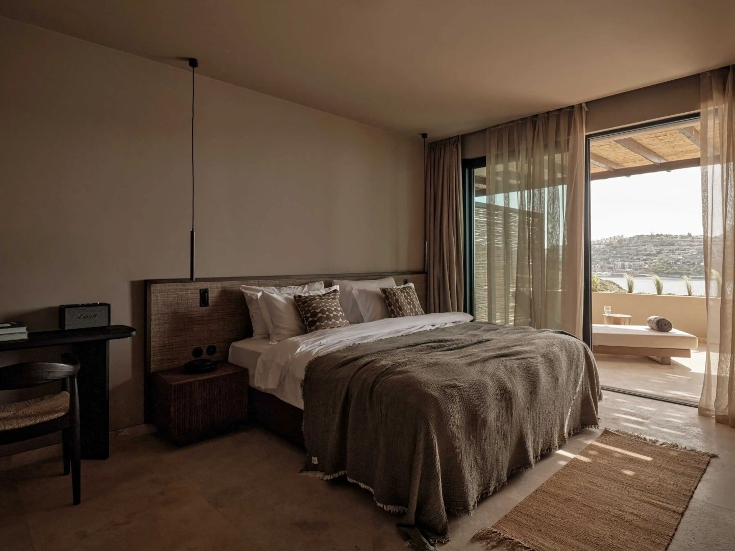 Doppelbett mit brauner Tagesdecke und Blick auf die angrenzende Terrasse mit Meerblick.