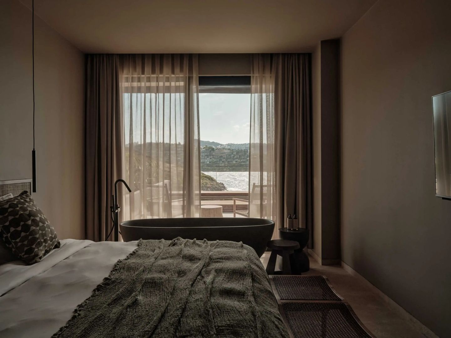 Schlafzimmer mit Doppelbett, freistehender Badewanne und Blick durch die Glasfront auf das Meer.