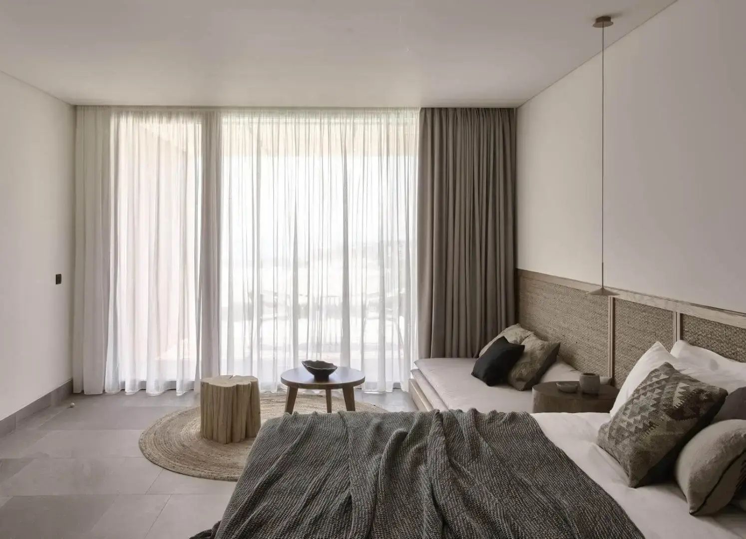 Sitzbereich im Hotelzimmer mit Holztisch, rundem Teppich und bodentiefen Fenstern mit Vorhängen.