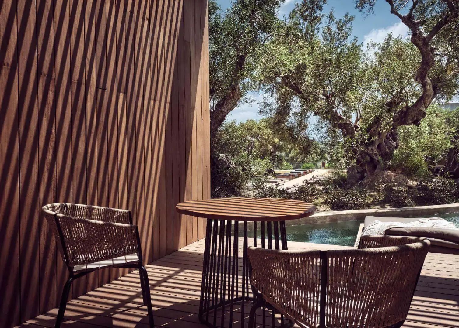 Sitzbereich auf einer Holzterrasse im Schatten neben Olivenbäumen und Wasser.