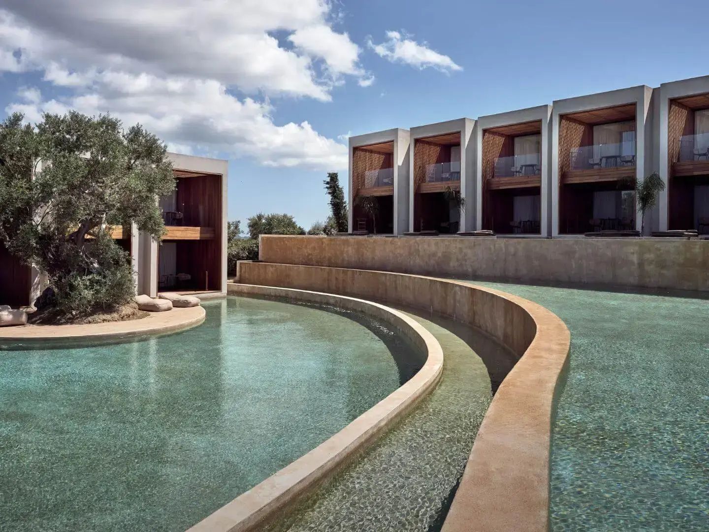 Geschwungene Poollandschaft vor modernen Hotelgebäuden aus Beton und Holz.