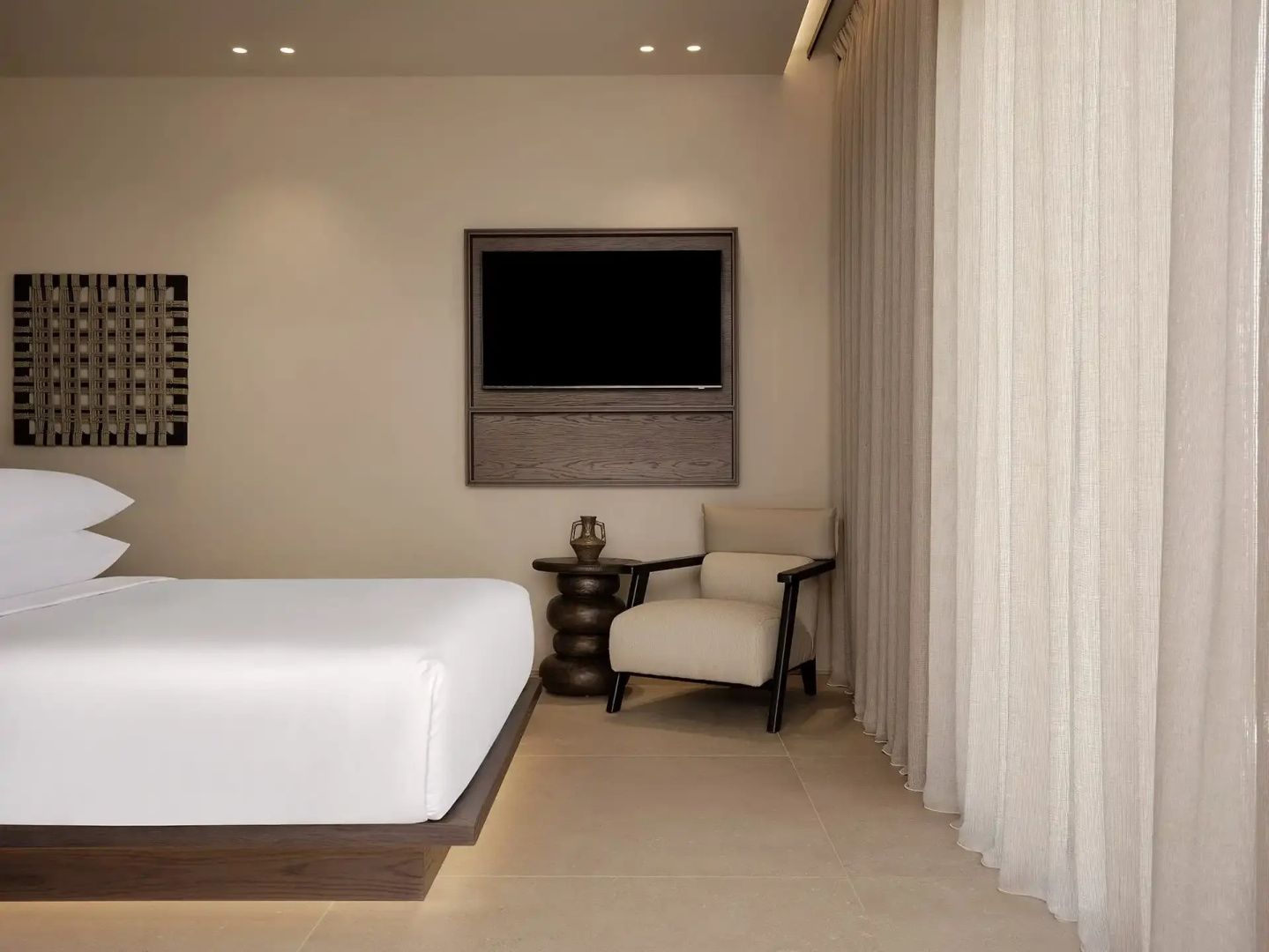 Hotelzimmer mit weißem Bett, wandmontiertem Fernseher und Sessel vor hellem Vorhang.