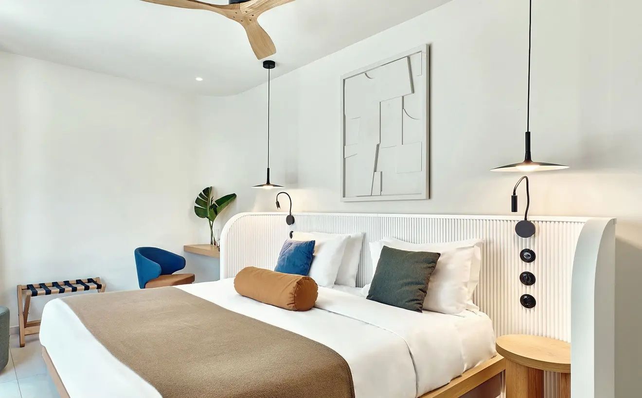 Doppelbett mit brauner Decke, Kissen, Holzventilator und blauem Sessel im hellen Zimmer.