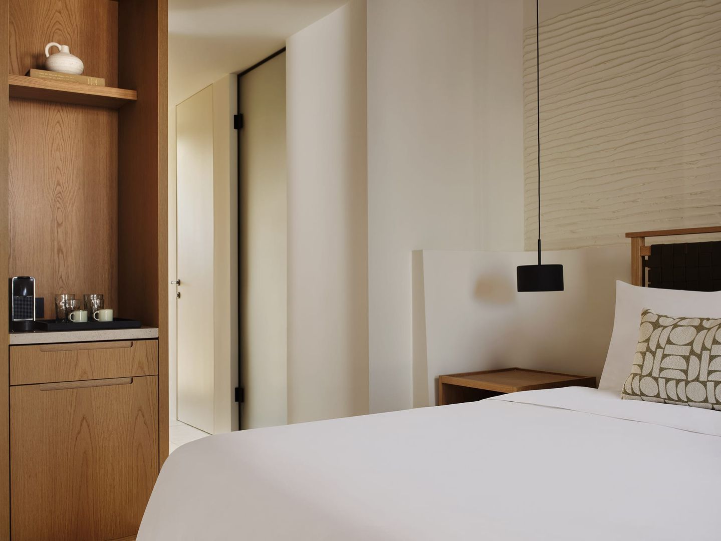 Helles Schlafzimmer mit weiß bezogenem Bett, Holzschrank und schwarzer Hängeleuchte.