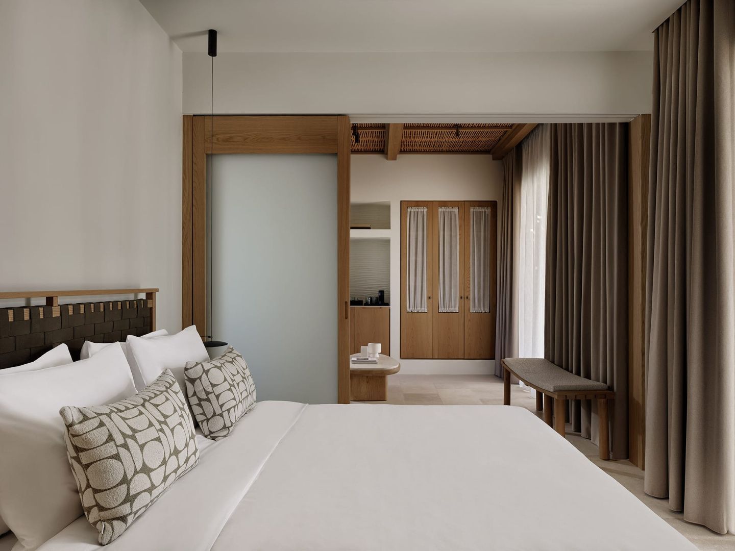 Helles Schlafzimmer mit Doppelbett, gemusterten Kissen und Holzschiebetür zum Wohnbereich.