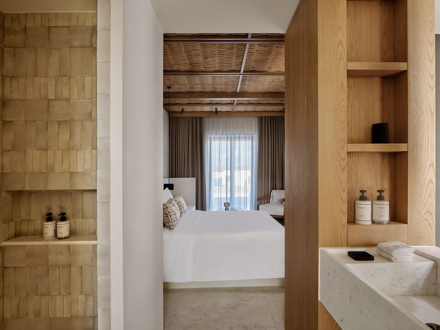 Blick in ein helles Hotelzimmer mit Holzbalkendecke, Doppelbett und heller Fensterfront.
