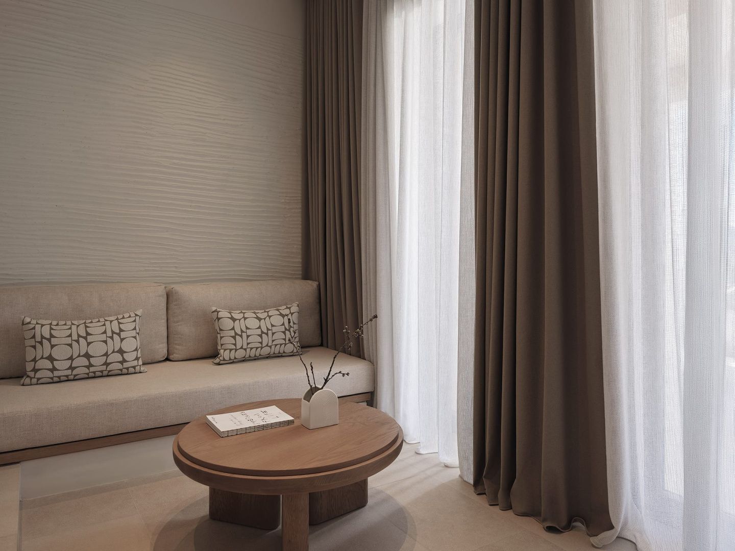Sitzecke im Hotelzimmer mit hellbrauner Couch, rundem Holztisch und bodentiefen Vorhängen.