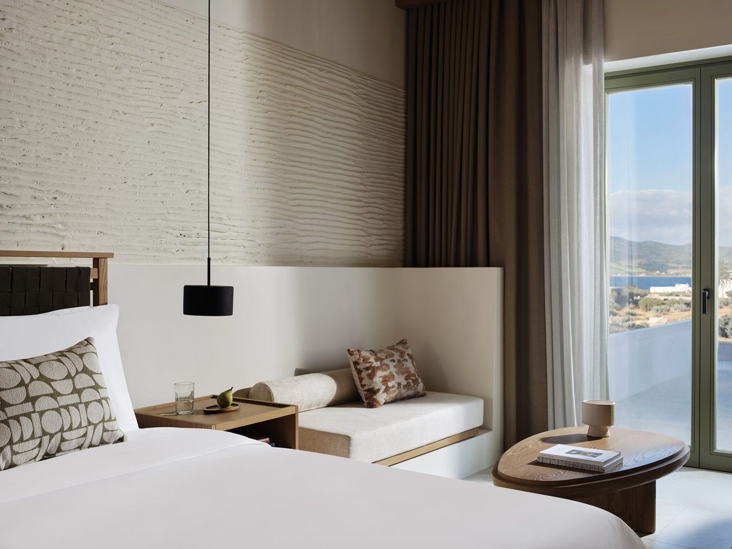 Helles Hotelzimmer mit Doppelbett, Sitznische, Nachttisch und Blick ins Freie.