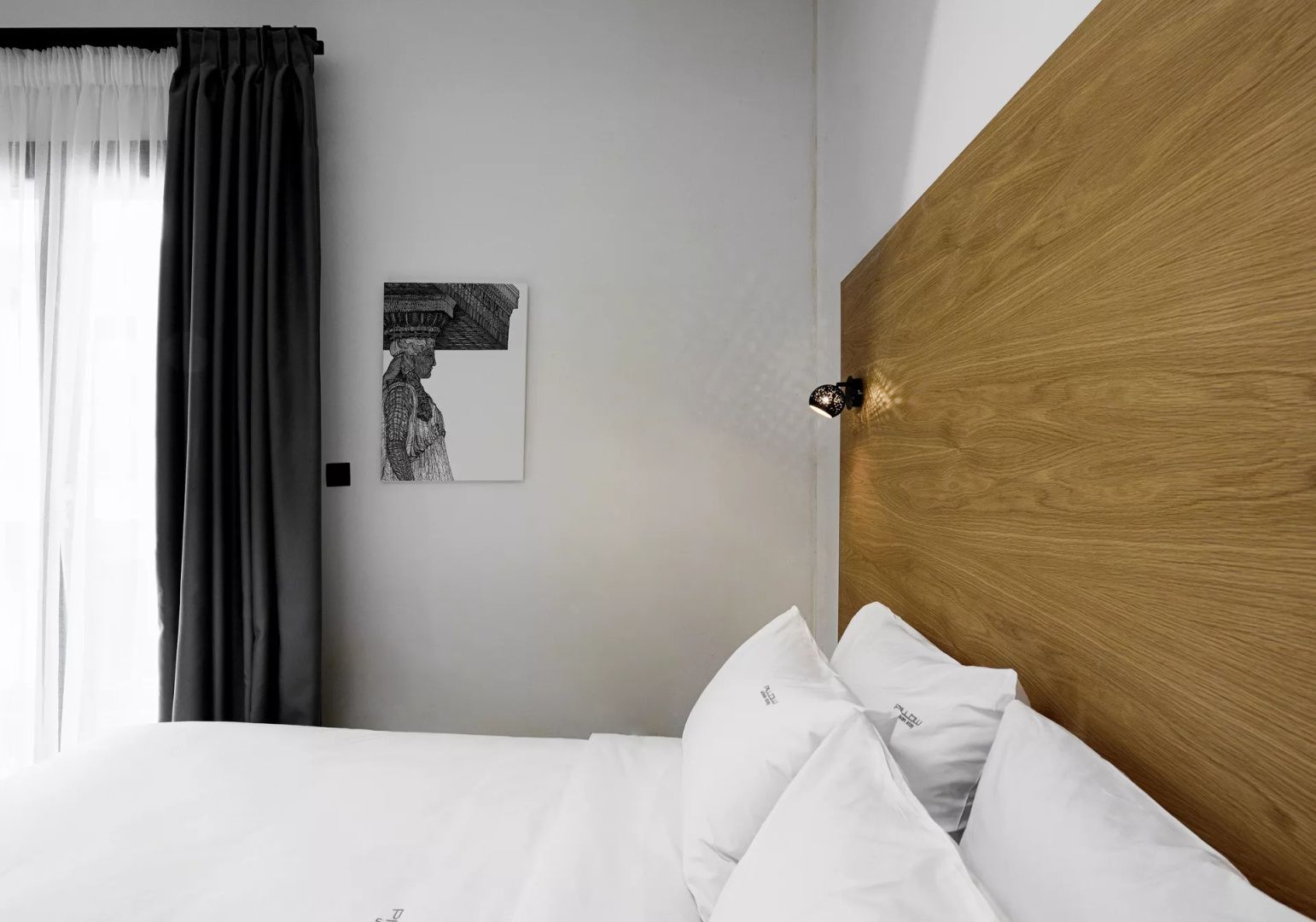 Nahaufnahme eines Doppelbetts mit Holzrückwand, weißer Bettwäsche und dunklem Vorhang.