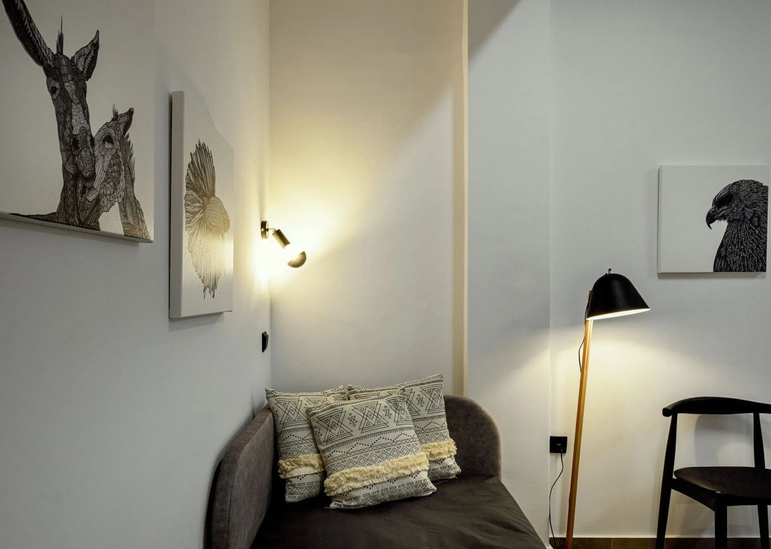 Sitzecke mit grauer Polsterbank, gemusterten Kissen, Wandleuchte und einer Stehlampe.