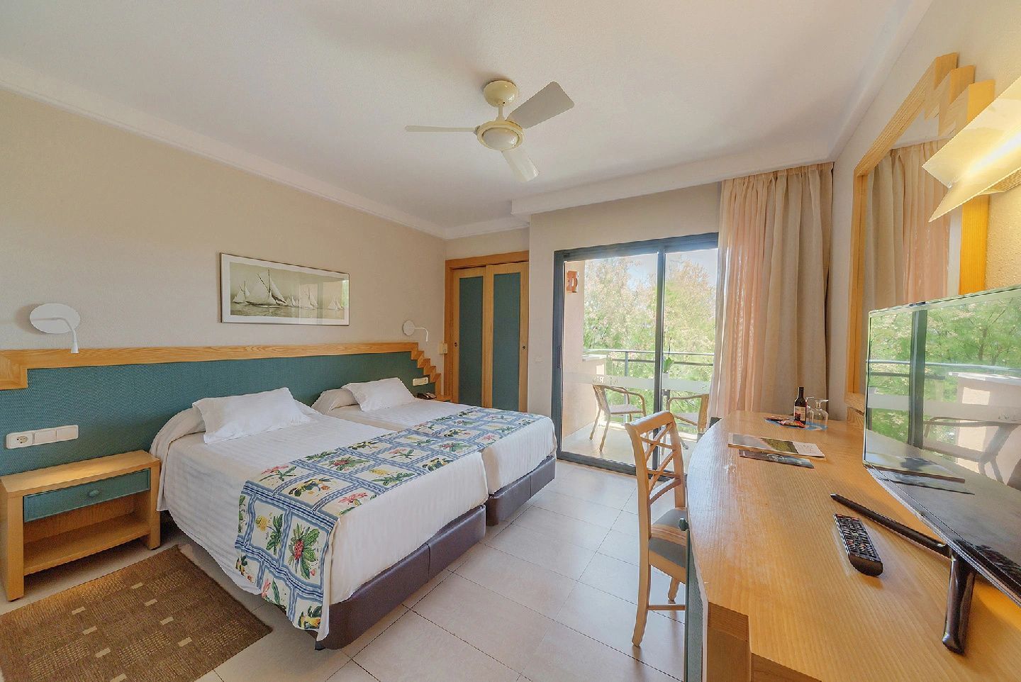 Helles Hotelzimmer mit Einzelbetten, Schreibtisch, Deckenventilator und Zugang zum Balkon.