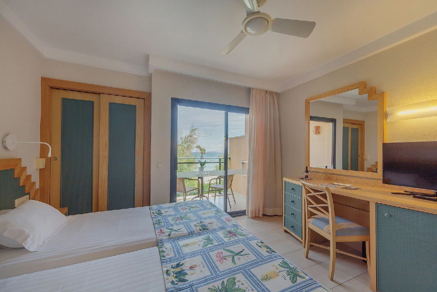 Doppelzimmer mit Deckenventilator, Holzmöbeln und großer Glastür zum Balkon.