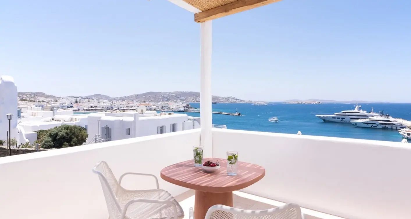 Terrasse mit hölzernem Tisch und zwei Stühlen mit Blick auf das Meer und weiße Häuser.