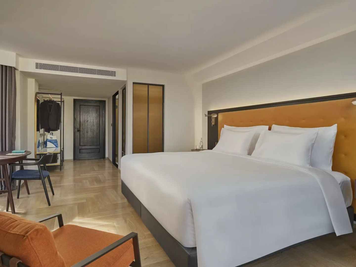 Modernes Hotelzimmer mit weiß bezogenem Doppelbett, orangefarbenem Sessel und Parkettboden.