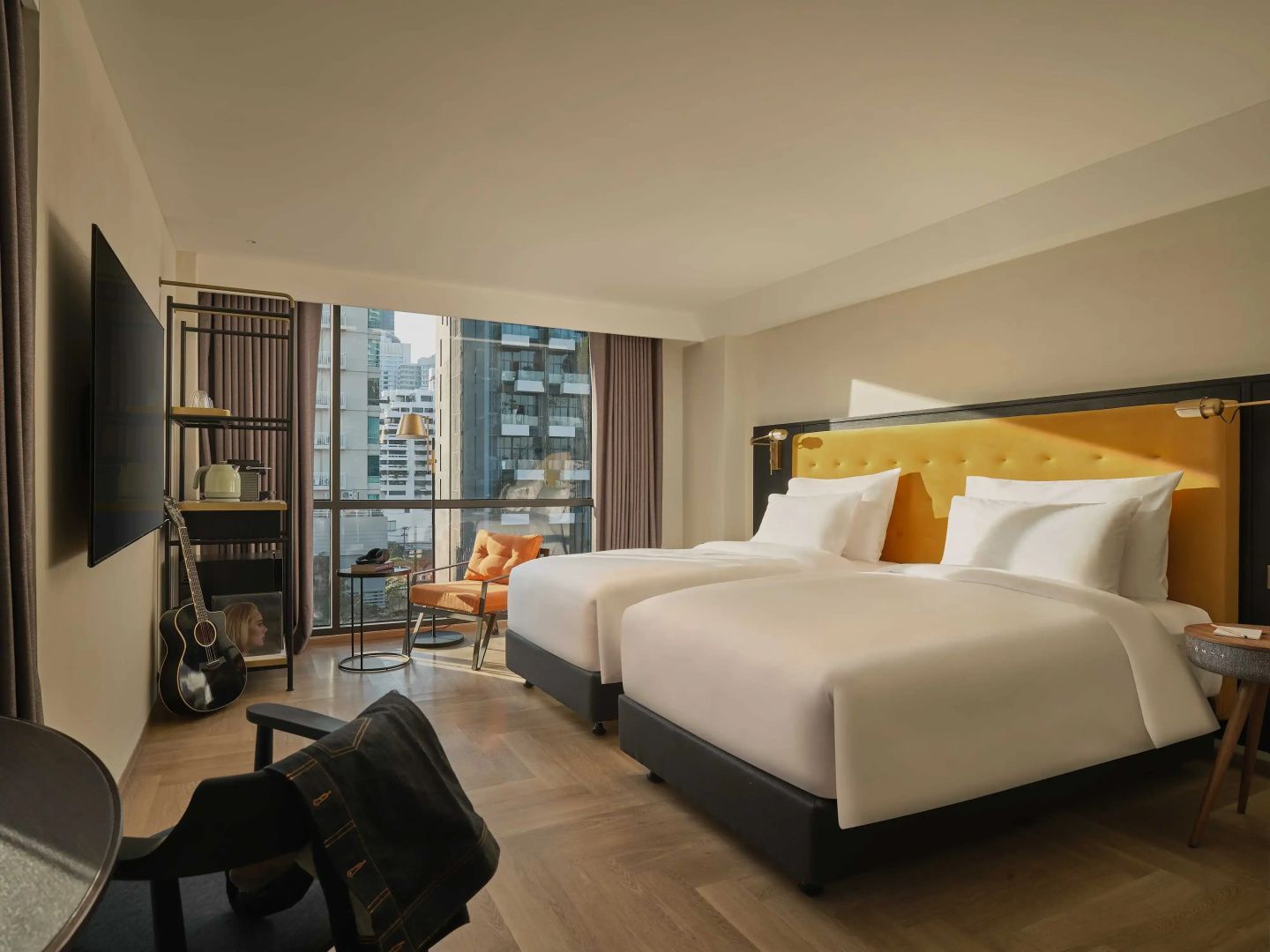 Hotelzimmer mit zwei Einzelbetten, gelbem Kopfteil, Sessel und großem Fenster mit Stadtblick.