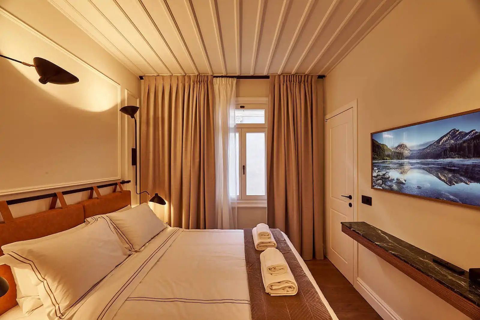 Hotelzimmer mit Doppelbett, beigefarbenen Vorhängen und Fernseher an der Wand.