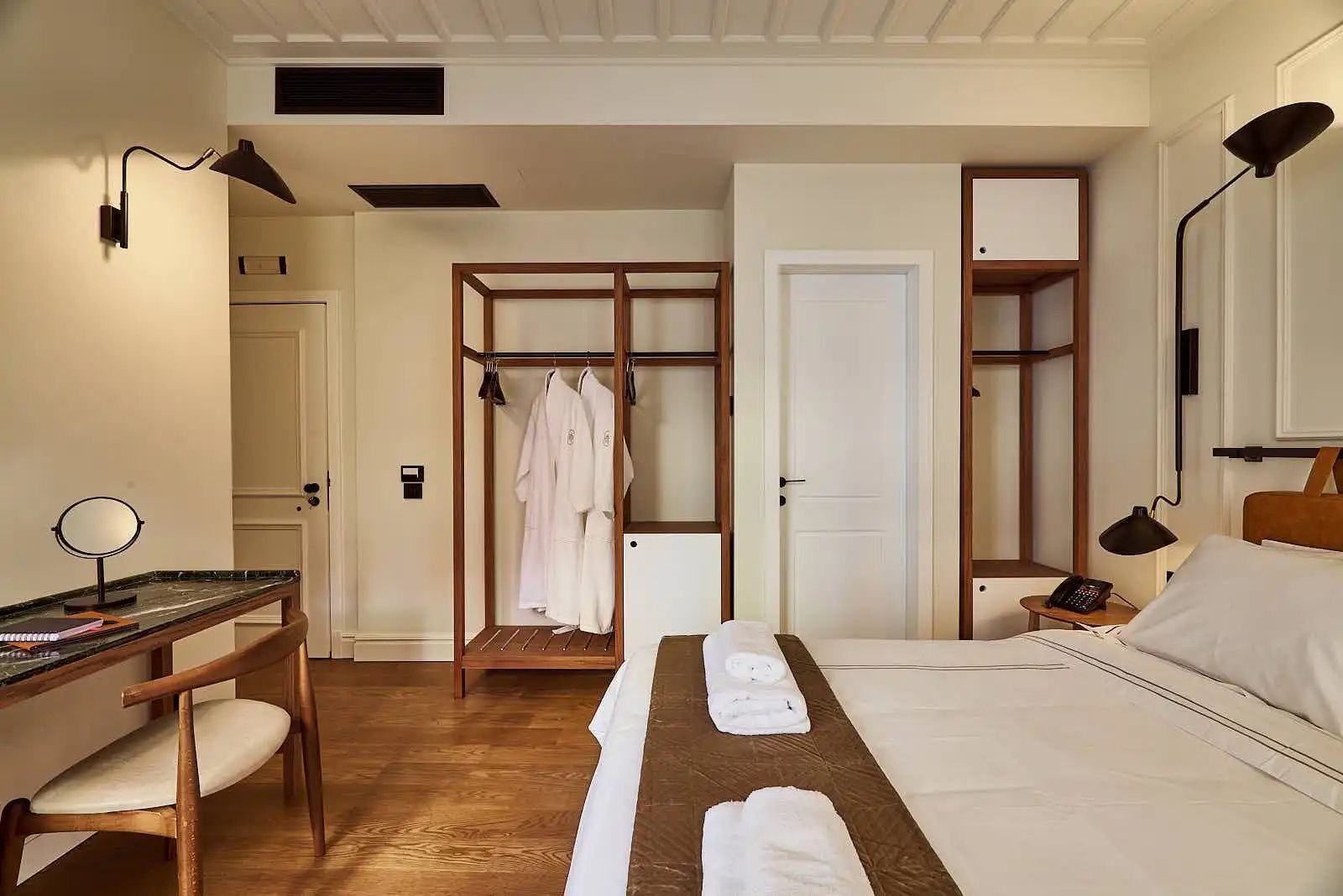 Helles Hotelzimmer mit offenem Holzregal, weißem Bademantel und Bettwäsche.
