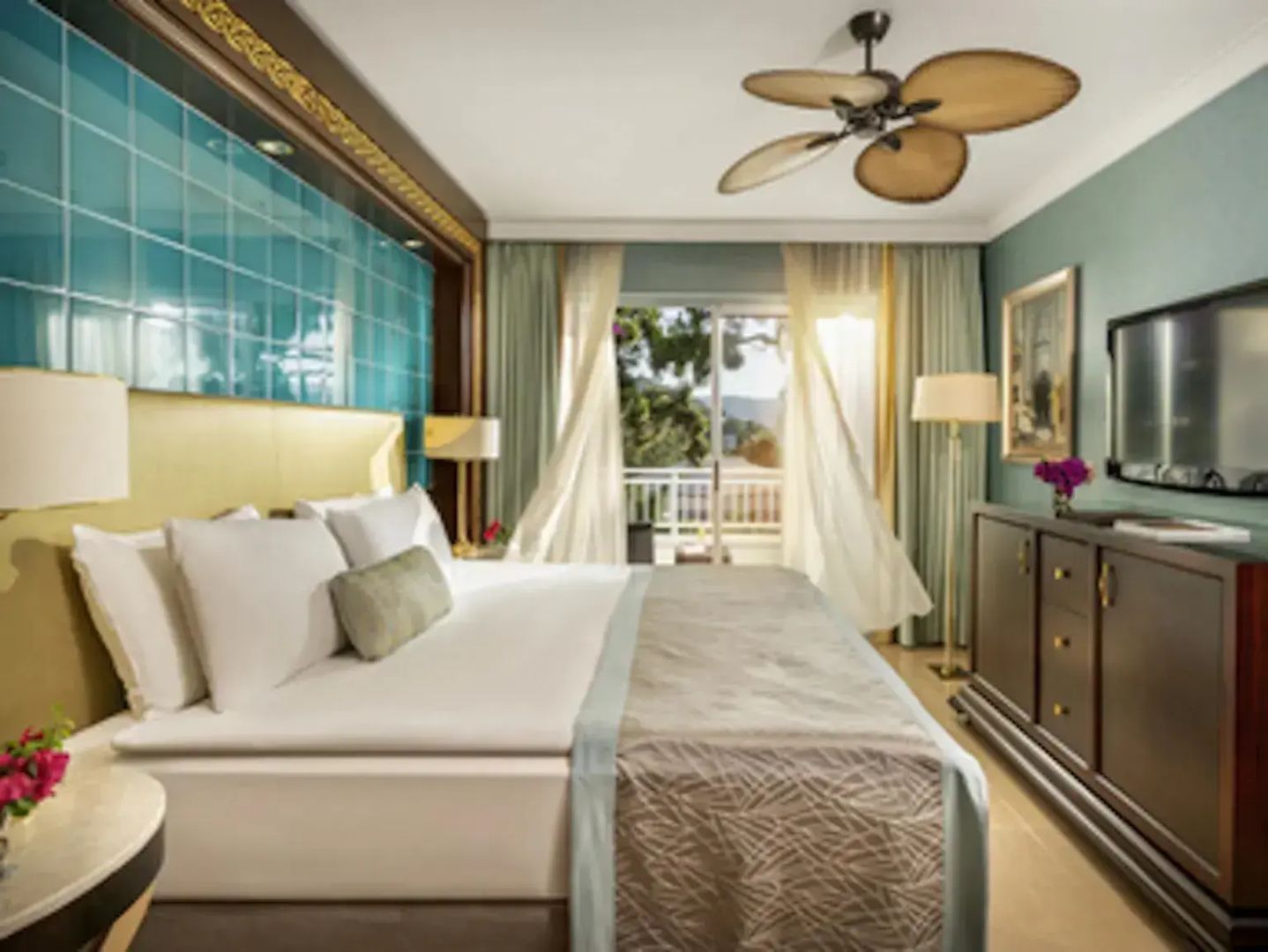 Hell gestaltetes Hotelzimmer mit großem Doppelbett, Deckenventilator und Vorhängen am Balkon.