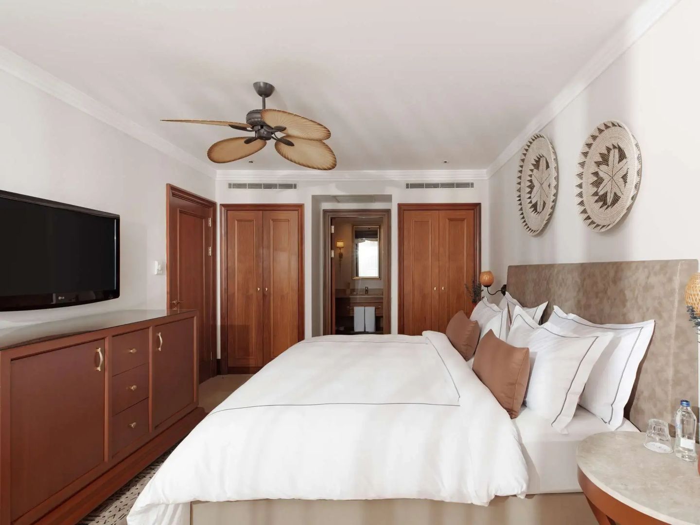 Helles Schlafzimmer mit Doppelbett, Holzschränken, Deckenventilator und Blick ins Badezimmer.
