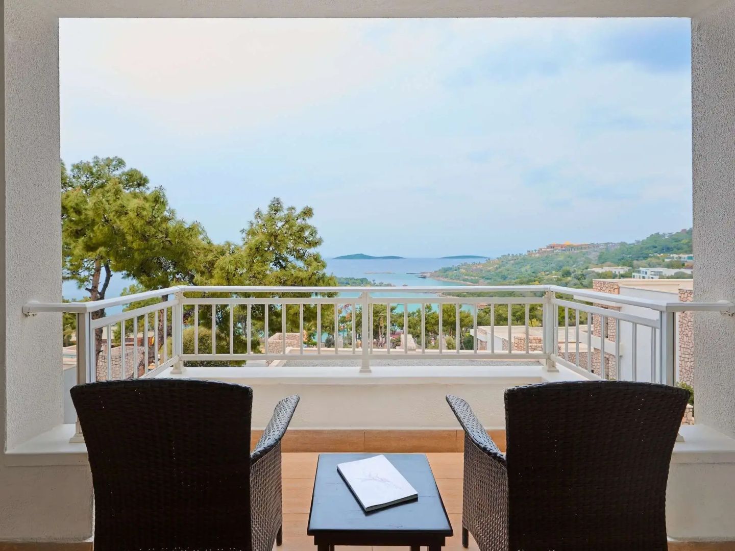 Blick vom Balkon mit zwei Stühlen auf das Meer und umliegende grüne Hügel.