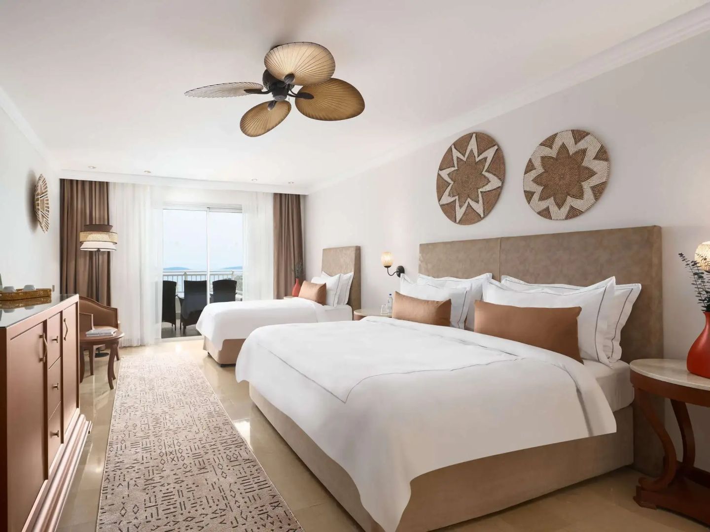Helles Hotelzimmer mit Doppelbett, Einzelbett, Deckenventilator und Balkonzugang.