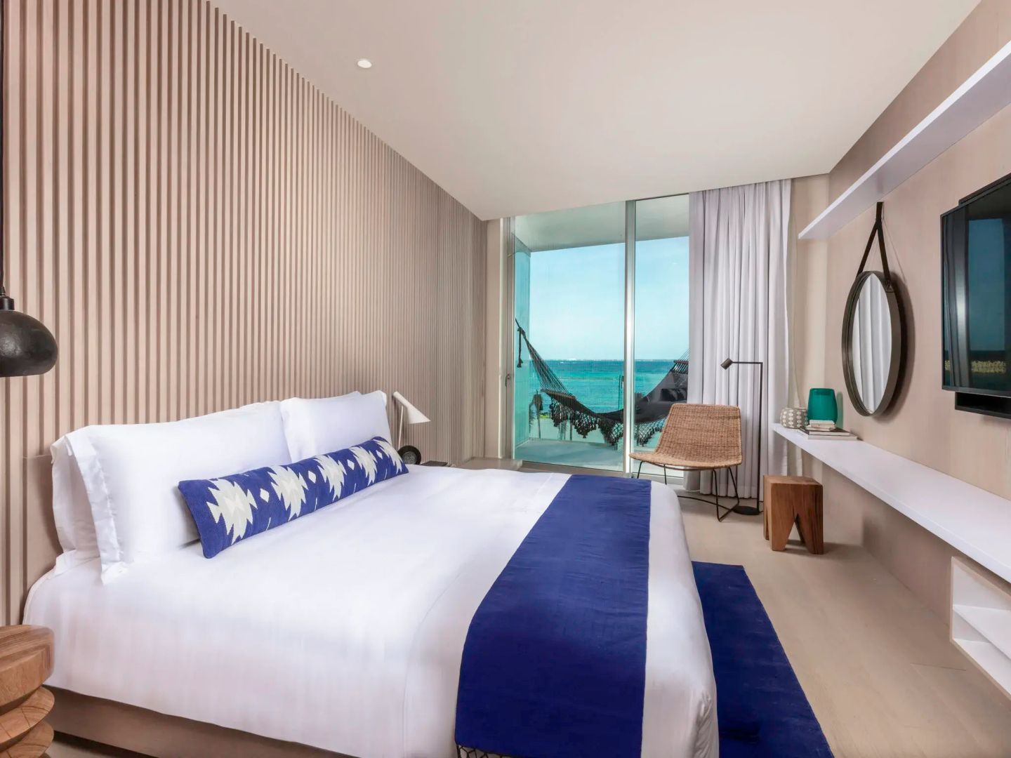 Helles Hotelzimmer mit Doppelbett, blauer Tagesdecke und Blick durch das Fenster auf das Meer.
