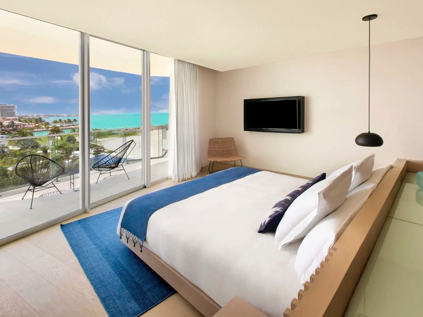 Doppelbett im hellen Hotelzimmer mit Fernseher und Blick durch große Fenster auf das Meer