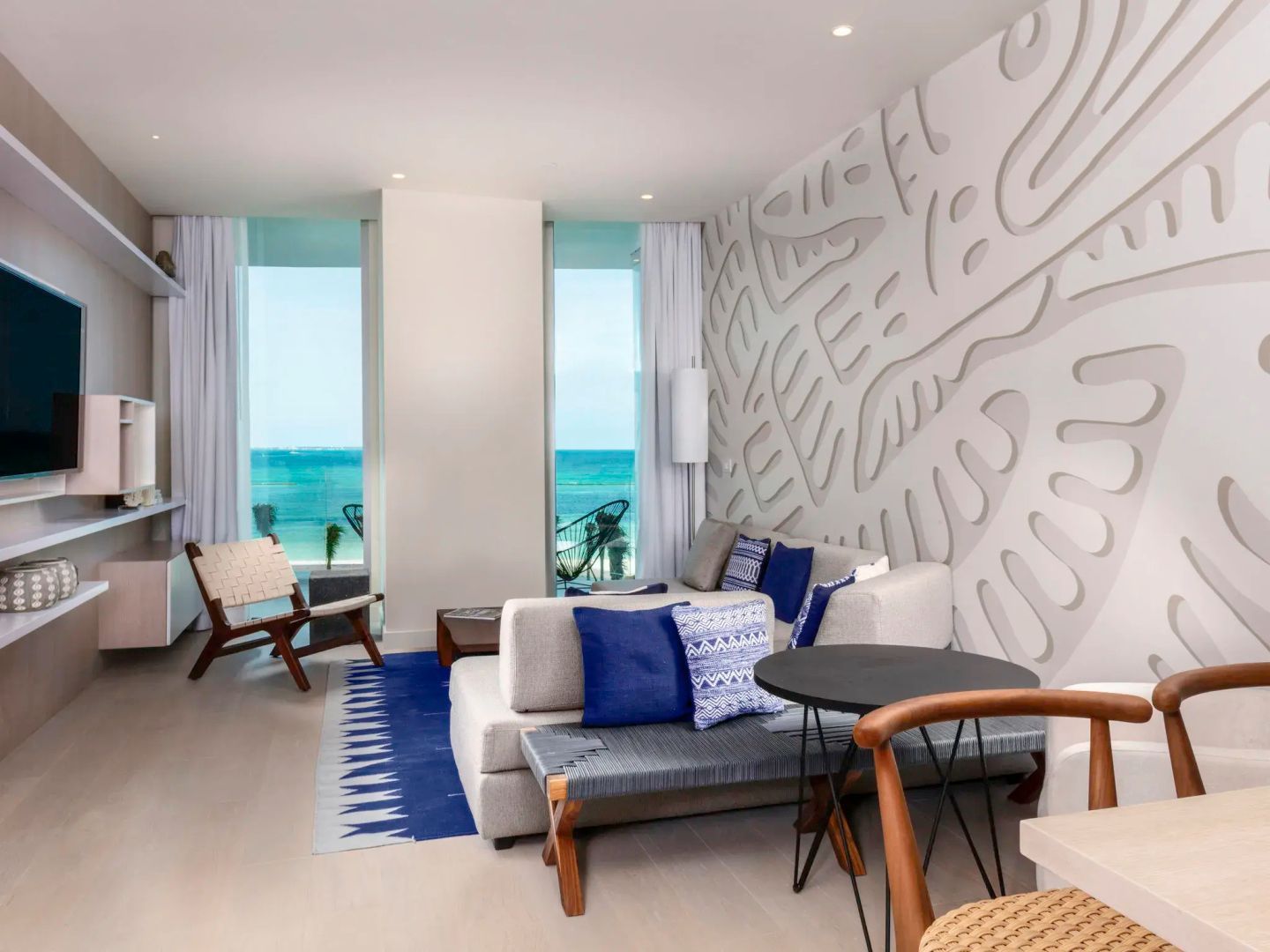 Sitzecke in einer Suite mit graustufiger Couch, blauen Kissen und Fensterfront zum Meer.