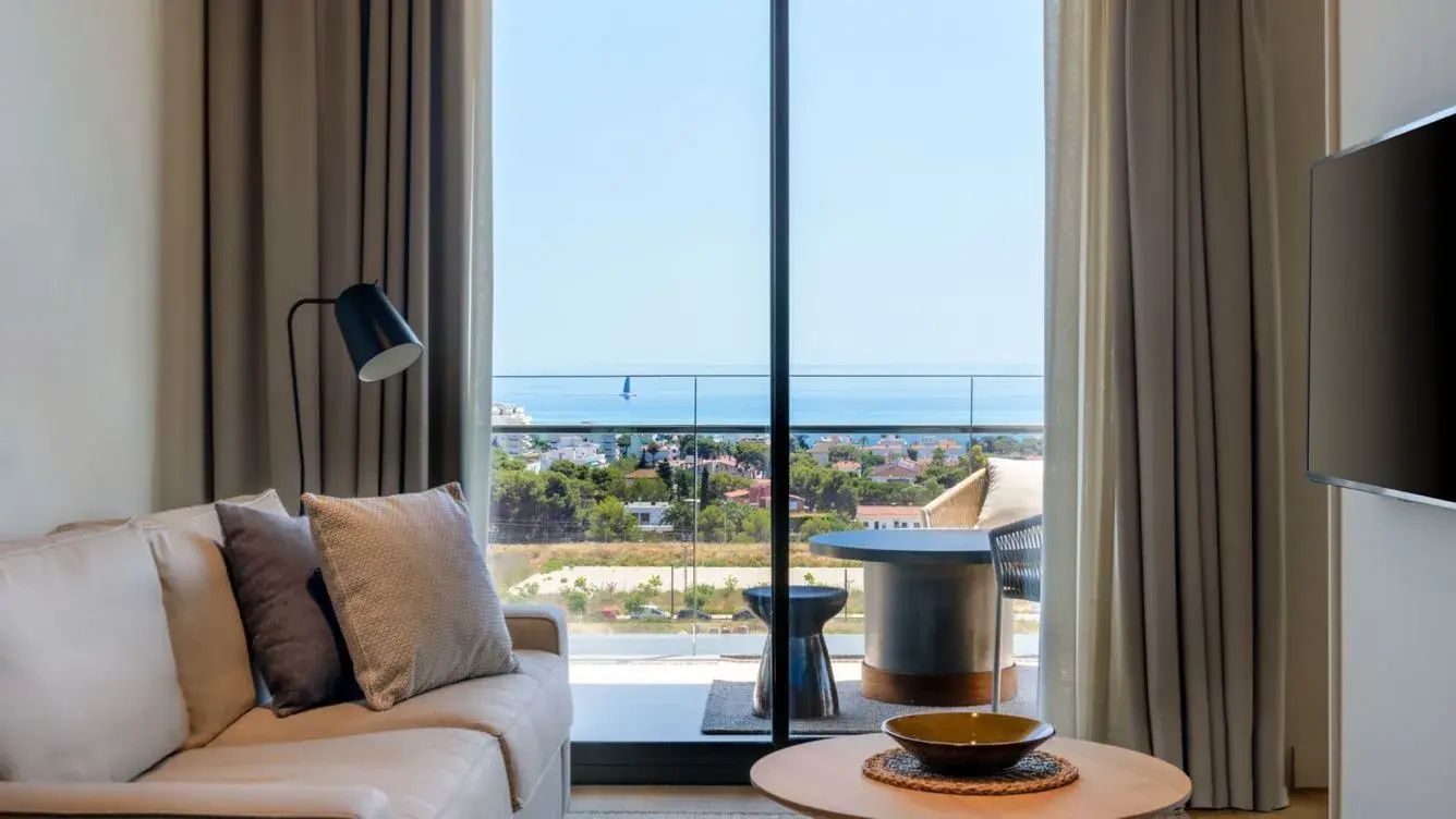 Ausblick vom Zimmer über den Balkon hinweg auf die grüne Umgebung und das blaue Meer am Horizont.