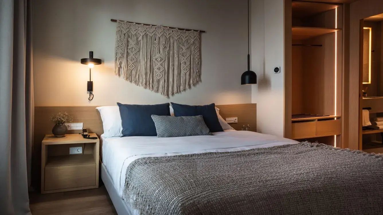 Doppelbett mit weißer Bettwäsche, blauen Kissen und Macramé-Wandbehang an einer hellen Wand.