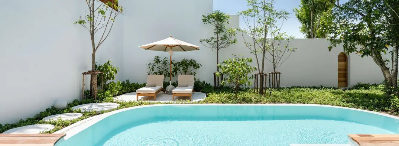 Privater Poolbereich mit zwei Sonnenliegen, Sonnenschirm und Begrünung vor weißer Wand.