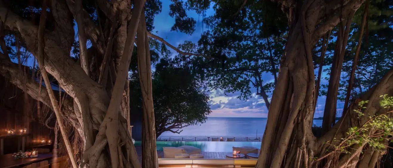 Blick bei Dämmerung über einen Pool auf das Meer, umgeben von Bäumen und Sitzmöglichkeiten.