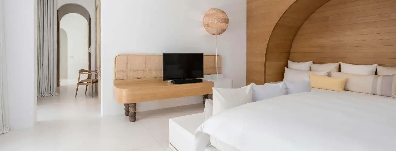 Schlafbereich mit weißem Bett, Holzsideboard mit Fernseher und einer Stehlampe.