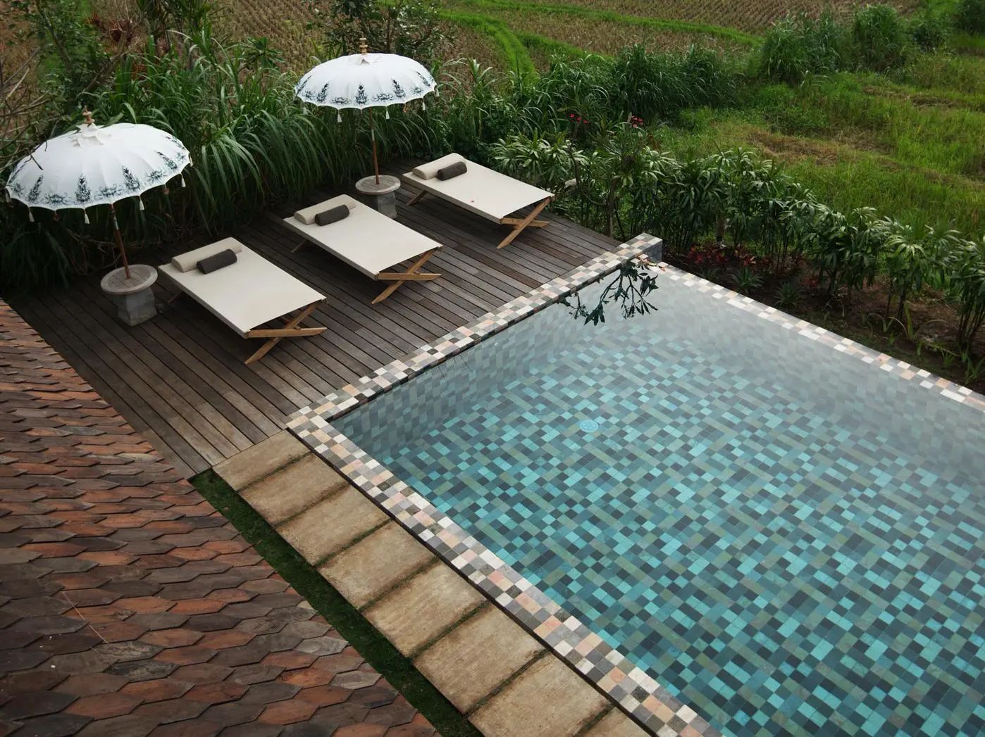 Swimmingpool mit türkisblauen Fliesen, Holzdeck und drei Sonnenliegen vor tropischen Pflanzen.