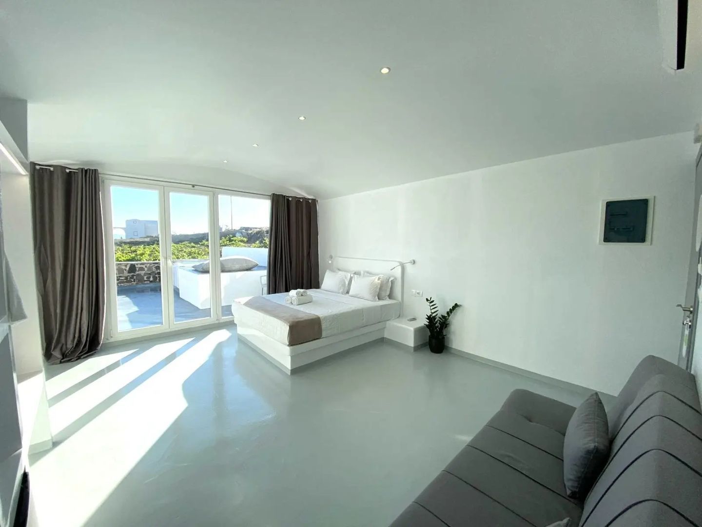 Geräumiges helles Zimmer mit Doppelbett, grauem Sofa und Glastür nach draussen.