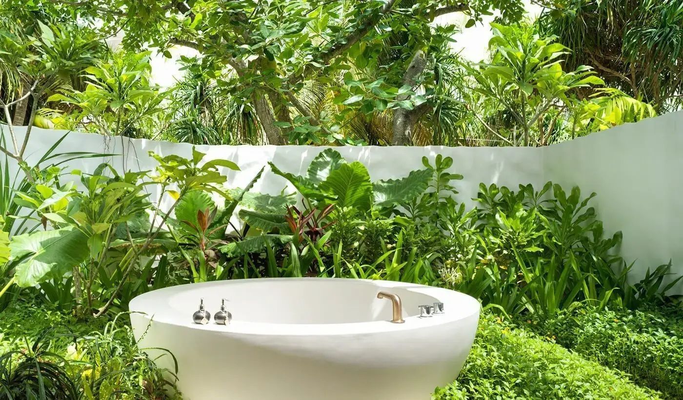 Freistehende weiße Badewanne im Außenbereich umgeben von tropischen Pflanzen.