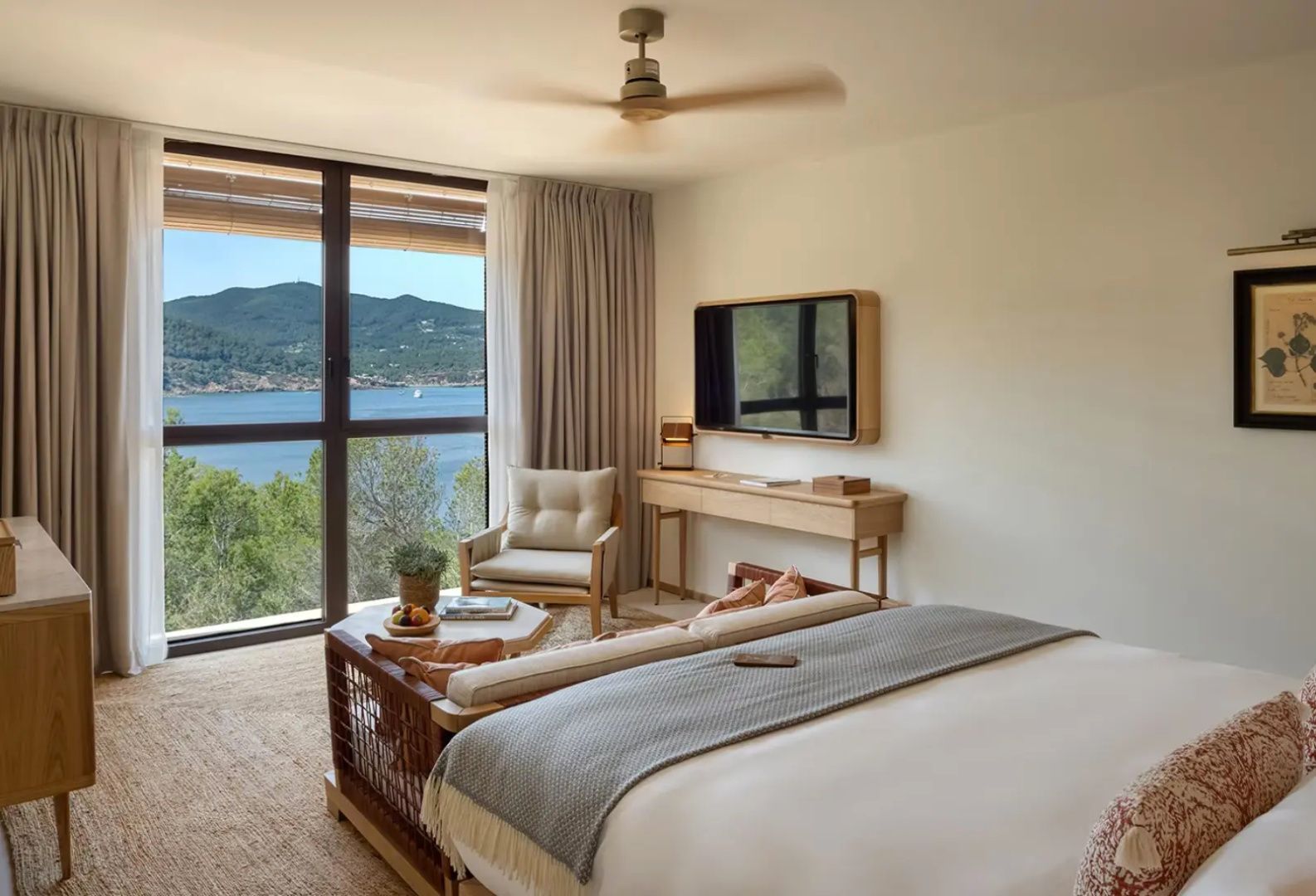 Helles Hotelzimmer mit Doppelbett, Sessel, Fernseher und Blick durch das Fenster auf das Meer.